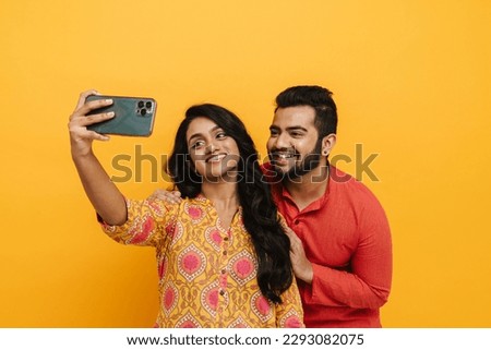 Cheerful beautiful indian couple taking selfie on smartphone isolated over yellow studio background