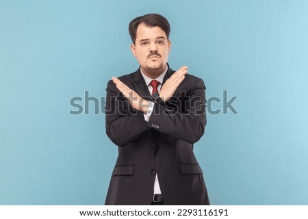 Portrait of angry upset man with mustache standing standing with crossed arms, saying no way, warning, wearing black suit with red tie. Indoor studio shot isolated on light blue background. Royalty-Free Stock Photo #2293116191