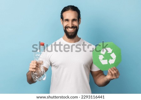 Portrait of responsible smiling man with beard wearing white T-shirt holding empty plastic bottle and green recycling sign, looking at camera. Indoor studio shot isolated on blue background.