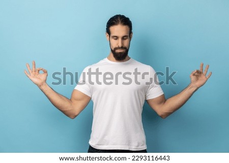Portrait of calm relaxed man with beard wearing white T-shirt doing yoga, breathing, keeping eyes closed, calming down after work. Indoor studio shot isolated on blue background. Royalty-Free Stock Photo #2293116443