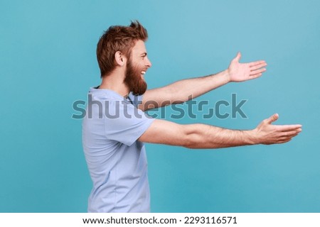 Free hugs, come into my arms. Side view of happy bearded man stretching hands to camera and smiling broadly, going to embrace, share love. Indoor studio shot isolated on blue background. Royalty-Free Stock Photo #2293116571