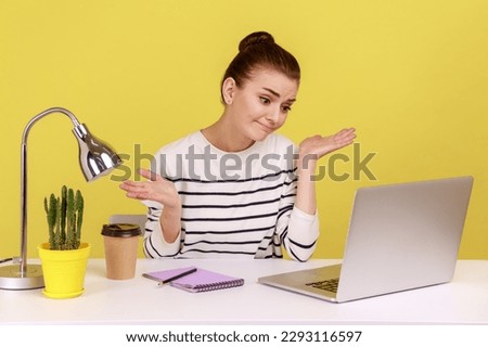 Uncertain woman office worker in striped shirt shrugging shoulders sitting on laptop at workplace, looking at monitor, having video call. Indoor studio studio shot isolated on yellow background. Royalty-Free Stock Photo #2293116597