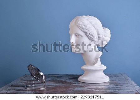 The concept of the psychology of self-esteem, beauty, self-care. A beautiful plaster figure of Aphrodite looks at herself in a small mirror, on a vintage wooden table. Royalty-Free Stock Photo #2293117511