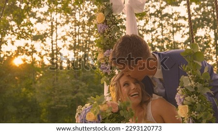 LENS FLARE: Handsome groom kisses his newlywed wife, who is sitting on a swing. Smiling newlyweds show each other affection while having a romantic moment in golden sunset light on their wedding day. Royalty-Free Stock Photo #2293120773
