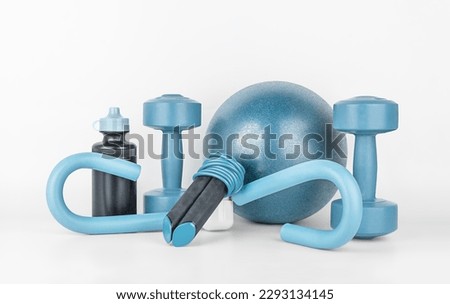 Fitness equipment on a gray background, front view, close-up. Dumbbells, sports bottle of water, jump rope, s-shaped leg exercise machine, fitness ball. Home workout. Fitness and activity.