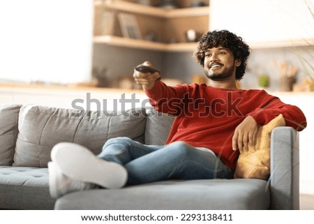 Home Relax. Handsome Indian Guy Sitting On Couch And Switching Tv Channels, Smiling Young Eastern Man Holding Remote Controller, Relaxing On Comfortable Sofa In Living Room, Copy Space Royalty-Free Stock Photo #2293138411