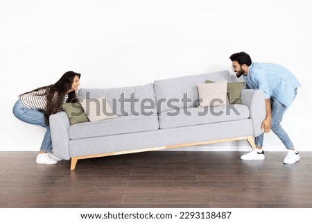 Happy excited beautiful indian young couple moving grey sofa in room over white wall background at new home, full length, copy space. Relocation, moving to new apartment concept