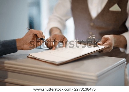 Cropped Shot Of Man Signing Papers Checking In At Hotel Reception Indoors. Male Staff Showing To Sign Documents Meeting Guest, Closeup Of Hands. Travel Accomodation Concept. Selective Focus Royalty-Free Stock Photo #2293138495
