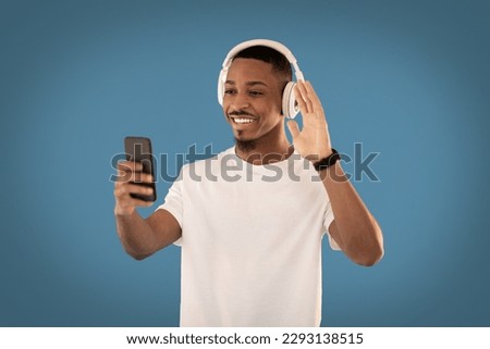 Telecommunication concept. Cheerful handsome young black guy in white t-shirt using brand new wireless headset and smartphone on blue studio background, have video call with friends Royalty-Free Stock Photo #2293138515
