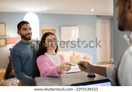 Cheerful Couple Checking In At Hotel Giving Credit Card To Receptionist Man Paying For Accomodation Room Standing At Reception Counter Indoors. Selective Focus On Happy Guests Royalty-Free Stock Photo #2293138523