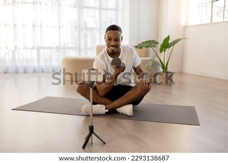 Handsome young black guy in sportswear fitness blogger streaming while exercising with barbell, happy sporty african man sitting on fitness mat at home, pumping iron and recording video on phone