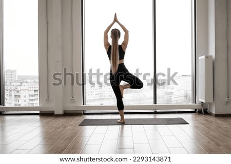 Back view of slender blonde woman in black top and yoga pants holding Tree Pose on rubber mat in meditation room. Attractive female calming mind in Vrikshasana exercise in front of panoramic window.