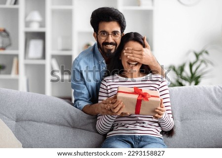 Loving young indian man covering woman eyes and giving gift box his beautiful wife, guy making great surprise for his girlfriend on birthday, home interior, free space. Anniversary celebration