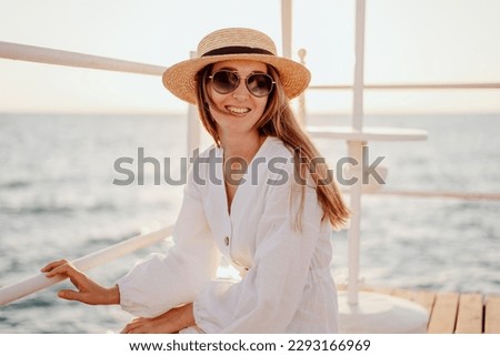 A beautiful young girl with long hair in a white dress, sunglasses and a straw hat smiles beautifully against the backdrop of the evening ocean.