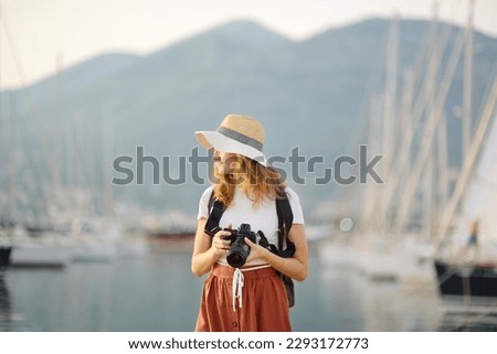 Charming young tourist woman taking a picture on the Mediterranean coast. Attractive red-haired girl photographer with a camera on the background of sea and yachts. Tourism and travel concept.