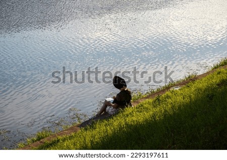 Back view of a young girl in hat sitting on the embankment of the river and drawing a sketch on a warm sunny summer day