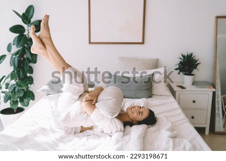 Side view of cheerful barefoot female with closed eyes relaxing on bed in spare time with pillow in folded hands and lifting crossed legs up Royalty-Free Stock Photo #2293198671