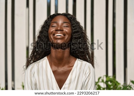 Happy cheerful young ethnic black African attractive pretty woman with curly long hair standing outdoor on sunny city street laughing. Close up portrait, candid authentic shot. Royalty-Free Stock Photo #2293206217