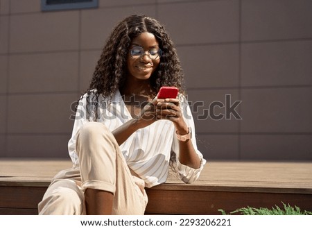 Young African black happy woman model holding cell phone using mobile apps for ecommerce shopping in online applications, communicating online on cellphone tech device sitting outside in city park.