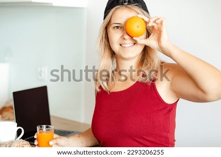 Young blonde in baseball cap is having fun at home in kitchen during breakfast with orange juice, puts orange to her eyes. Works online at home. Healthy eating. Positive emotions. Humor. 