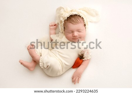 A cute little newborn baby in a white suit and a hat with rabbit ears sleeps sweetly. In the hands of a red felted carrot. Image of a rabbit, professional photo closeup on a white background.