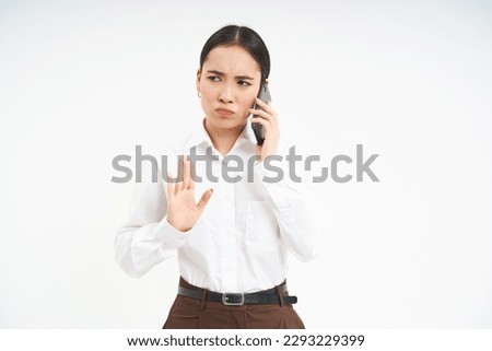 Portrait of serious korean woman, business professional shows stop sign, talks on mobile phone with disapproval, frowns upset, stands over white background.