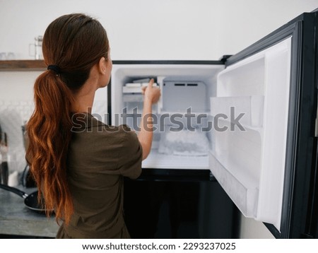 Woman smiling with teeth looking into camera in kitchen at home opened freezer empty with ice inside, home refrigerator, defrosted, view from back, stylish interior. Royalty-Free Stock Photo #2293237025