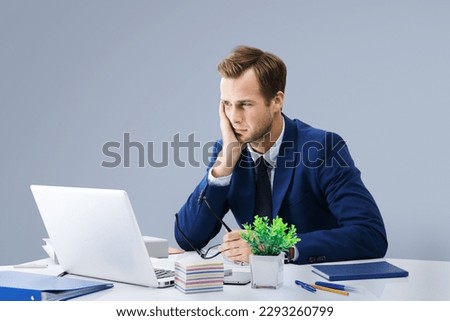 Stressed, tired businessman or heaving headache, holding his head, looking at laptop computer, office workplace. Confident man - business, job and education concept. Grey wall background. Royalty-Free Stock Photo #2293260799