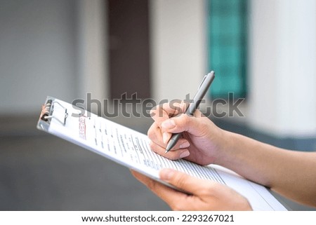 An engineer hand is using a pen checking on the building inspection report form to QC building quality of the house (background). Export industrial working scene concept. Close-up and selective focus. Royalty-Free Stock Photo #2293267021