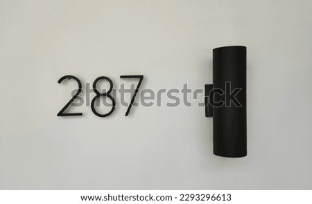 House number 287 in matt black is placed on a white wall and has a modern style lamp in matt black.