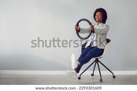 Funny, happy African American woman sitting on a chair in a room with a light gray copy space wall, holding a black steering wheel cover in her hands, smiling and pretending to drive an invisible car Royalty-Free Stock Photo #2293301609