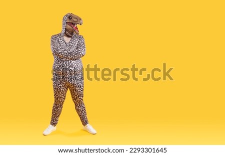 Funny eccentric overweight man posing in dinosaur mask on his head and in pajamas with leopard print. Bizarre fat man stands with crossed near copy space on orange background. Full length. Banner.