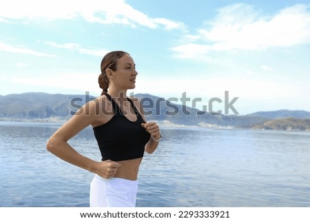 Fit woman wearing black top and white pants running on the promenade. Female model in a sportswear running along the coast on a beautiful summer day. Close up, copy space, background.
