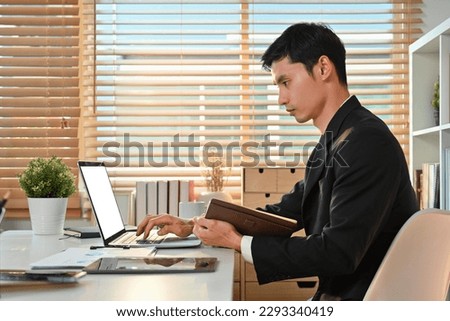 Side view of millennial asian male entrepreneur holding planner in hand and using laptop at bright office interior