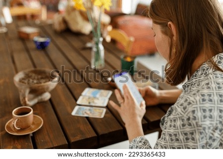 Beautiful young woman is guessing on cards with tarot, runes on wooden table and uses an online app in phone to interpret divination, home interior,candles and dry herbs.. Royalty-Free Stock Photo #2293365433