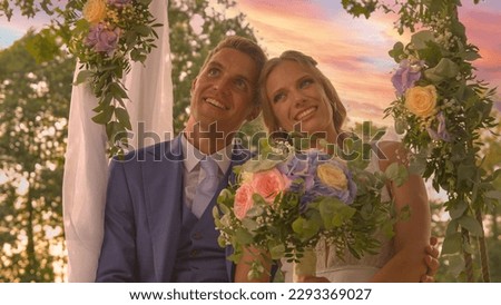 PORTRAIT, LENS FLARE: Smiling newlyweds posing on a beautifully decorated swing. Lovely bride and handsome groom in embrace of each other under colorful summer sunset clouds on their wedding day. Royalty-Free Stock Photo #2293369027
