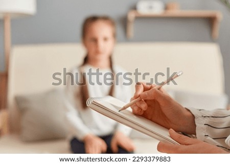 Closeup of doctor psychologist holding paper notebook making notes while talking with small girl at psychological therapy session meeting, blur defocused little kid patient. Royalty-Free Stock Photo #2293379883
