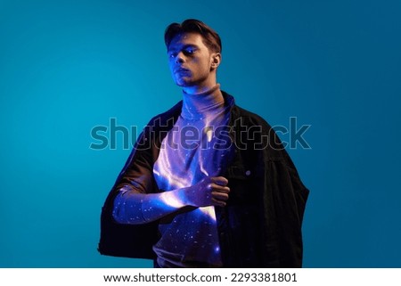 Handsome young man in stylish clothes with digital light reflection on body posing with serious face against blue background in neon light. Concept of modern photography, art, cyberpunk, creativity