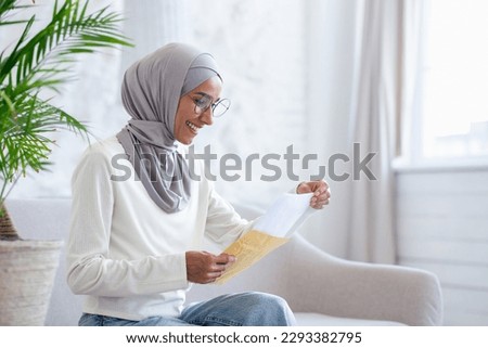 Joyful woman at home received letter mail notification with good news, Muslim woman in hijab is happy sitting on sofa in living room. Royalty-Free Stock Photo #2293382795