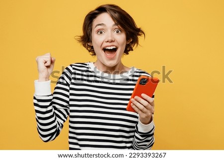 Young overjoyed happy fun woman wearing casual striped black and white shirt hold in hand use mobile cell phone in red case do winner gesture isolated on plain yellow color background studio portrait