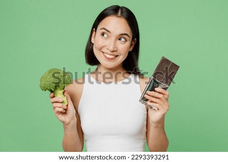 Young minded woman wears white clothes hold in hand dark chocolate bar broccoli vegetable isolated on plain pastel light green background. Proper nutrition healthy fast food unhealthy choice concept Royalty-Free Stock Photo #2293393291