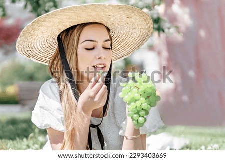 A young farmer woman eats white grapes picked by herself at the green grapes vineyard. Picnic in the blooming garden.
