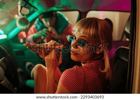 A retro fashionable girl is sitting in the car with her roller skates on and her boyfriend looking through the passenger window while she's eating a pizza slice and looking at the camera.