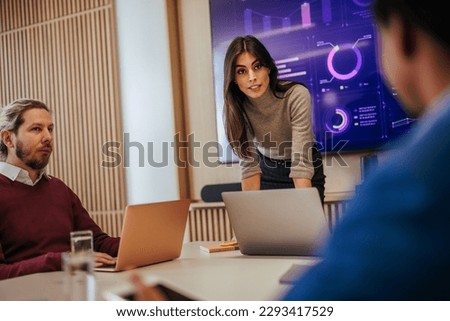A female team leader is with her coworkers in the meeting room working on development strategies on their laptops and with charts on the digital display on the wall. Royalty-Free Stock Photo #2293417529