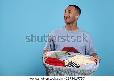 Happy man with basket full of laundry on light blue background. Space for text