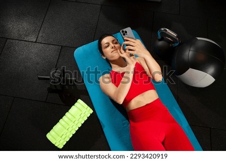 athletic woman in red sportswear lying on yoga matte and using smartphone in black gym, girl typing message on phone in fitness room, attractive woman in fitness club chatting online Royalty-Free Stock Photo #2293420919