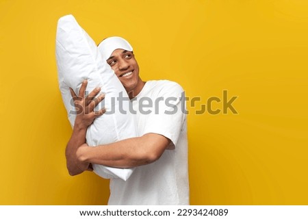 young african american guy in pajamas and sleep mask hugs soft pillow and dreams on yellow isolated background, man goes to bed and looks at the copy space, healthy sleep concept Royalty-Free Stock Photo #2293424089