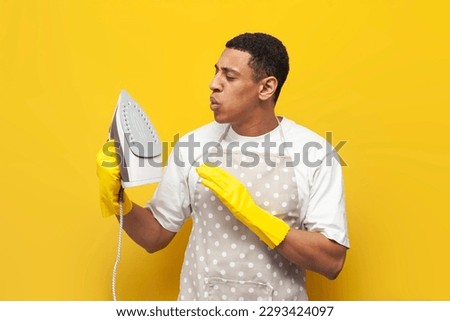 young guy african american cleaner in apron and gloves holds hot iron and blows on it on yellow isolated background