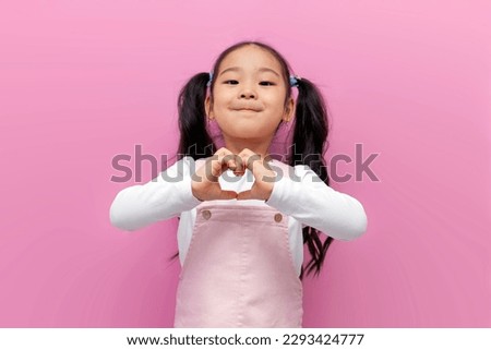little Asian girl in pink sundress with long hair shows heart with her hands, Korean preschool child makes gesture of love on pink isolated background