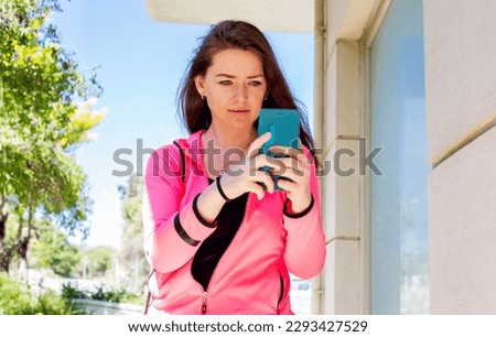 Beautiful young woman with long brown hair in pink sportswear using mobile phone taking selfie or making video call on the street